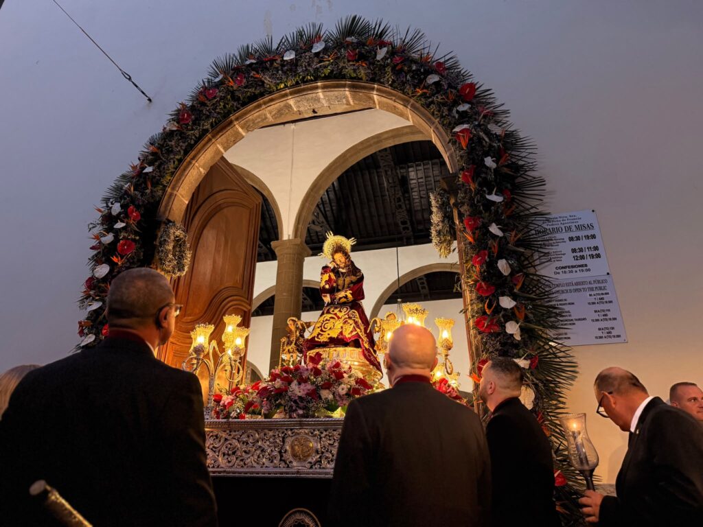 Gran Poder de Dios image carried beneath a flowered arch at the church entrance in July 2026