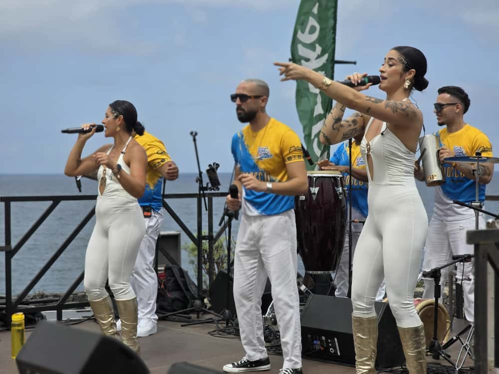 Singers and percussionists performing on a small stage by the sea at Summer Carnival 2025